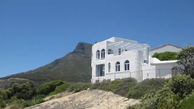 A large white house with arched windows stands on a rocky hillside beneath blue sky.