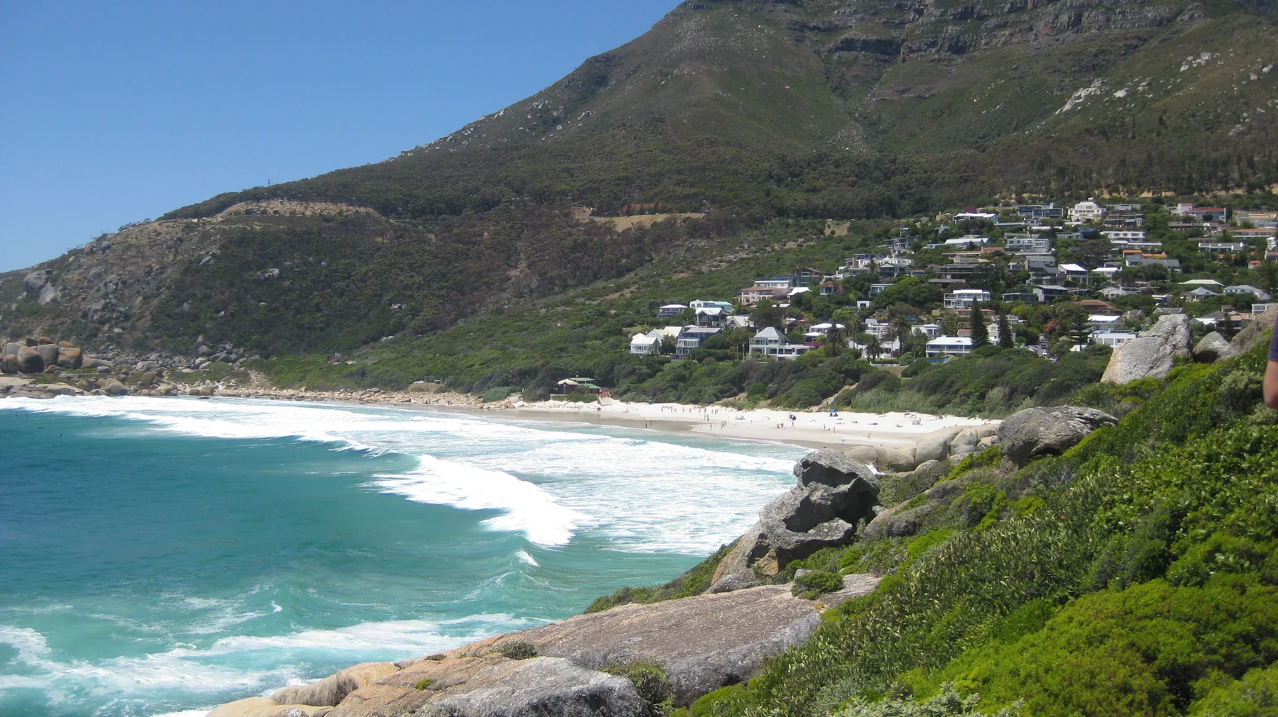 White houses dot a steep green hillside above a sandy cove and turquoise surf.