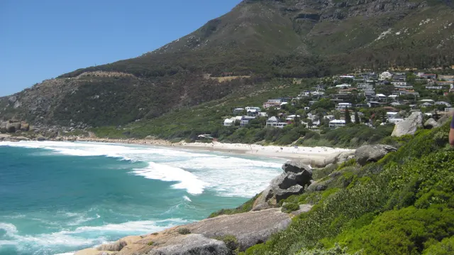 White houses dot a steep green hillside above a sandy cove and turquoise surf.