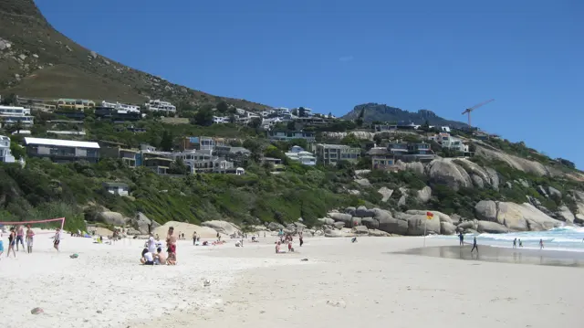 People gather on white sand below a hillside lined with white houses.