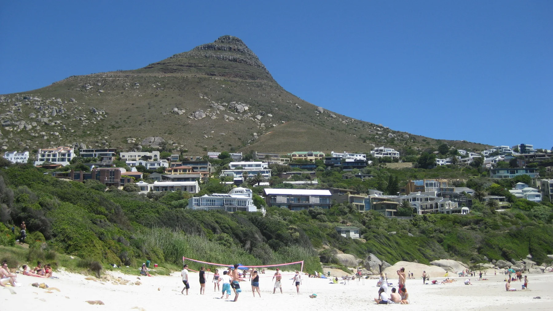 Beachgoers stand on pale sand beneath a steep conical peak and hillside homes.