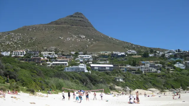 Beachgoers stand on pale sand beneath a steep conical peak and hillside homes.