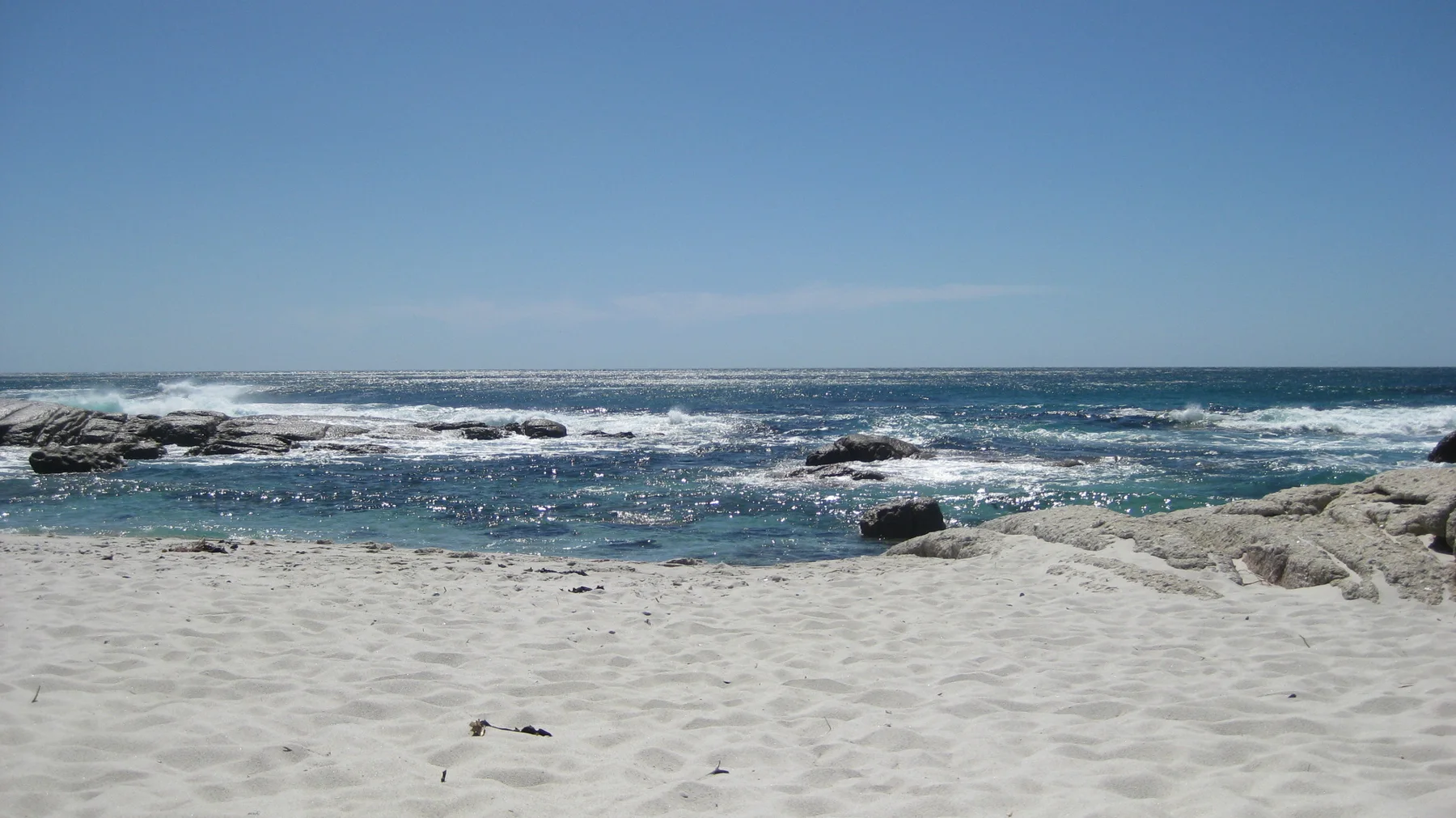 Dark rocks break the surface of blue shallows beyond a stretch of white sand.