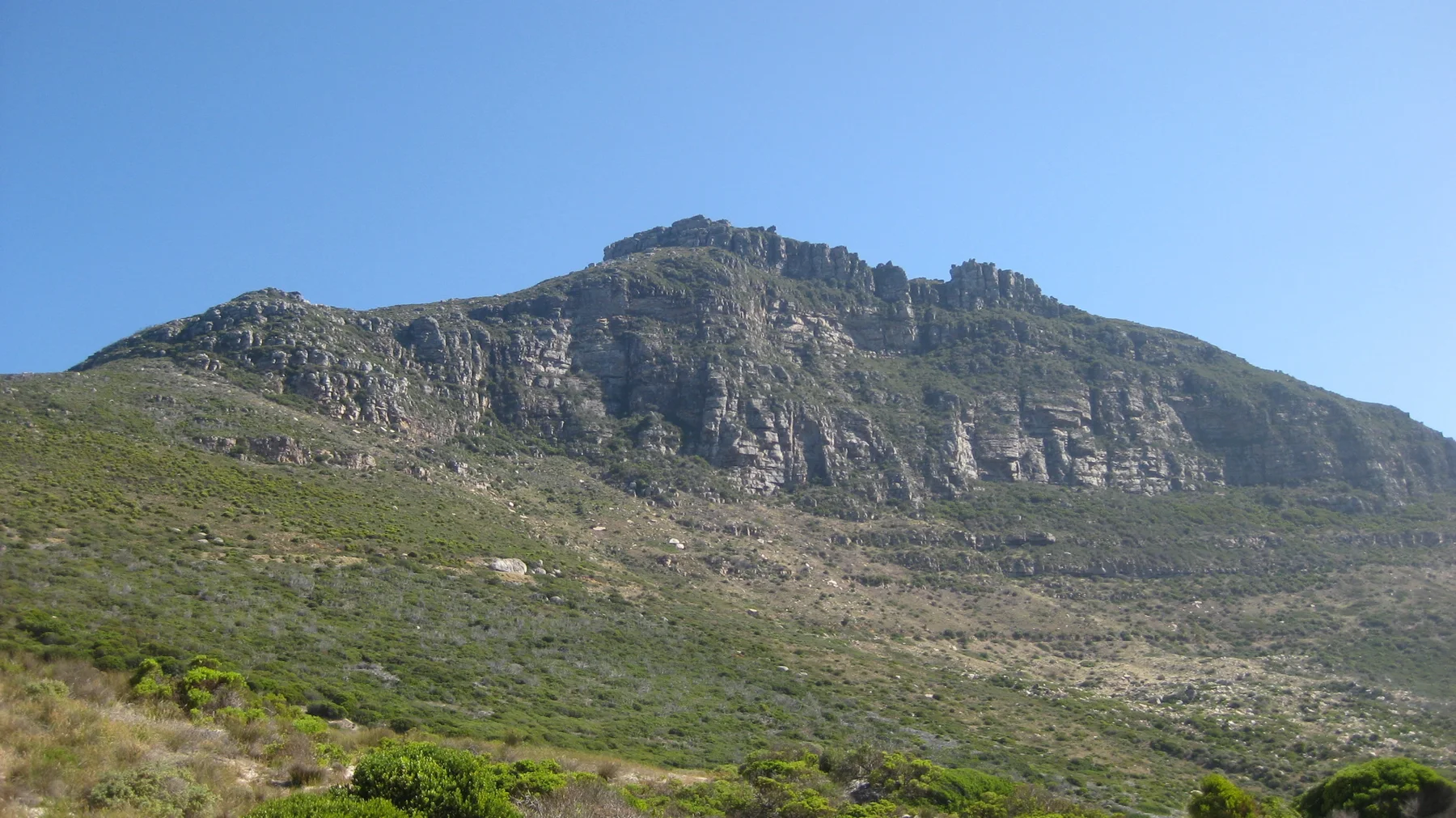 A broad rocky ridge rises above a green slope beneath clear blue sky.