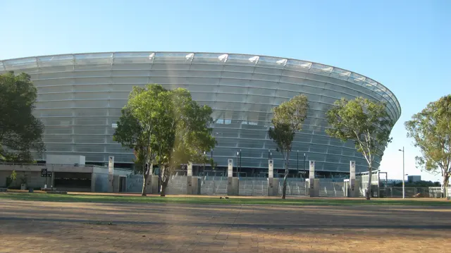 A large oval stadium with a silver facade rises behind trees and palms.