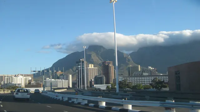 Traffic and city buildings fill the foreground beneath clouds spilling over a broad mountain.