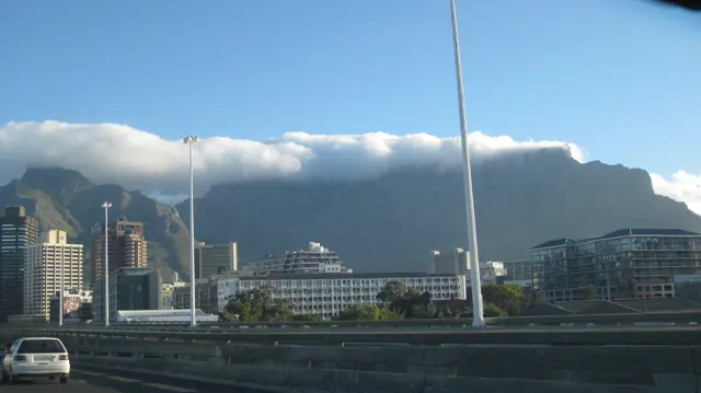 City buildings and a streetlight stand below a long mountain capped with cloud.