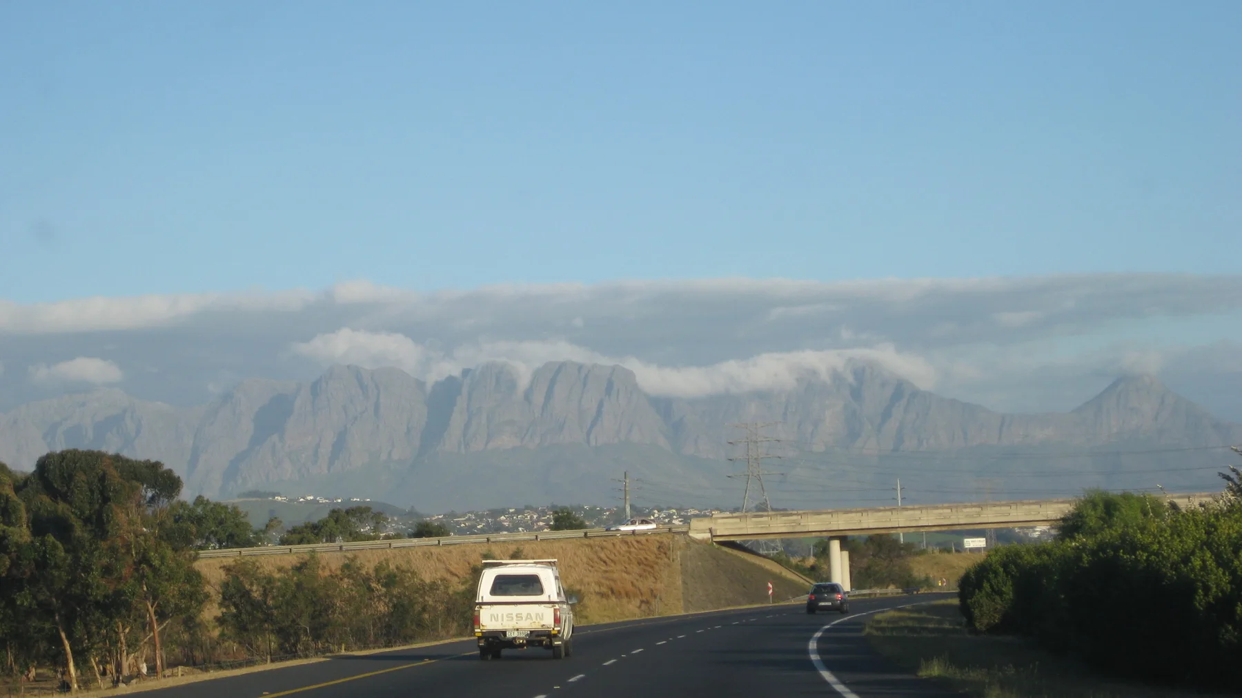 A closer view follows a white van along the road beneath a cloud-topped mountain.