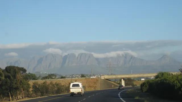 A closer view follows a white van along the road beneath a cloud-topped mountain.