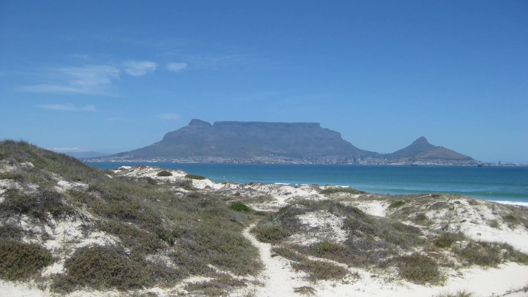 A narrow path crosses pale dunes and green vegetation toward turquoise water and a distant mountain.