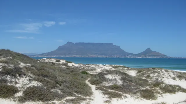 A narrow path crosses pale dunes and green vegetation toward turquoise water and a distant mountain.