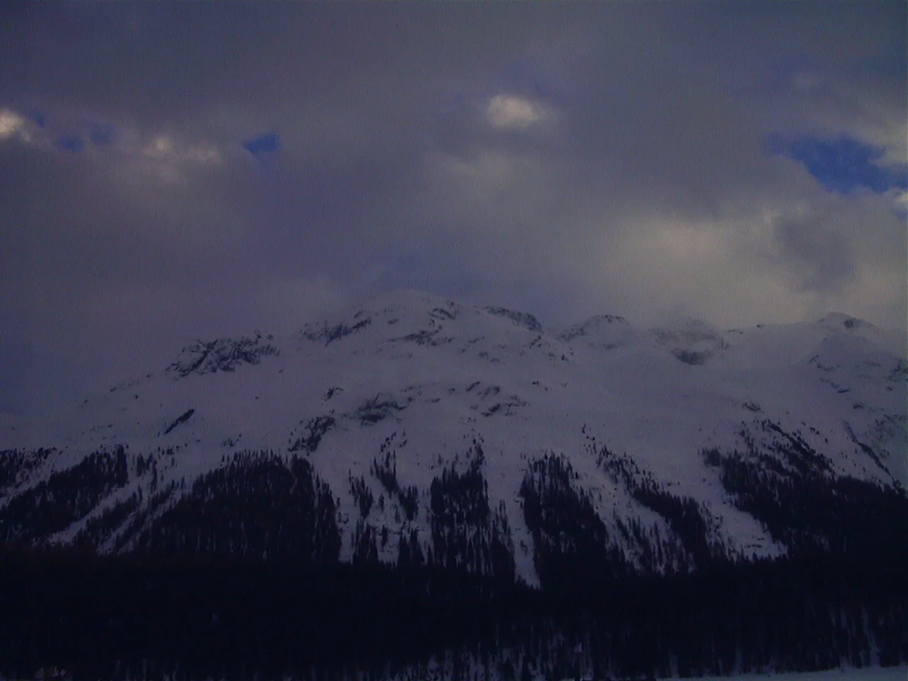 Snow-covered mountains rise above a dark forest beneath heavy clouds with small patches of blue sky.