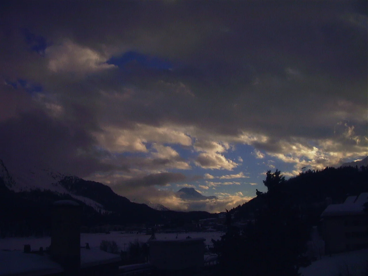 Warm light breaks through dark layered clouds above a shadowed winter valley.