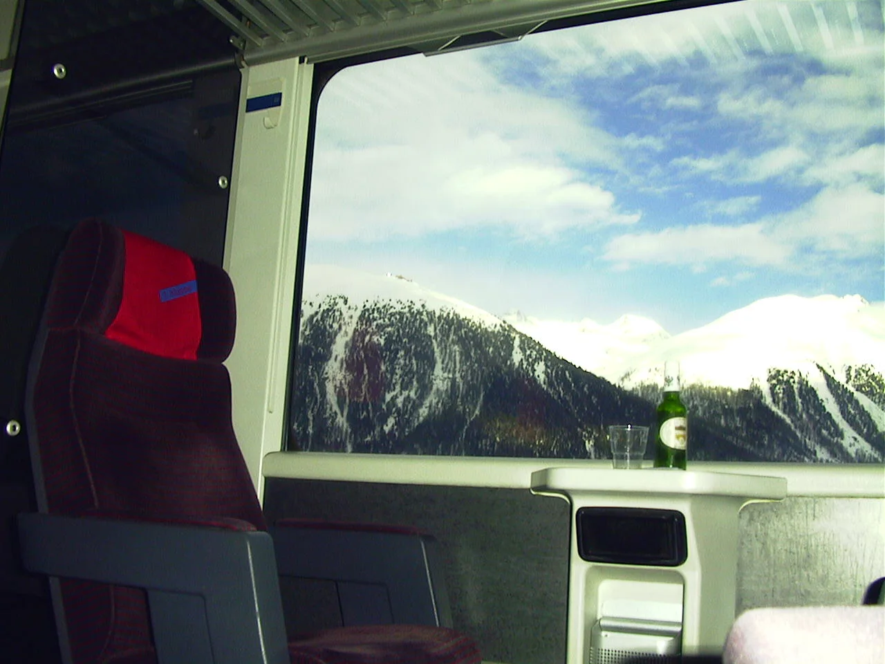 A red train seat and a small table frame a broad window looking towards snowy wooded slopes.
