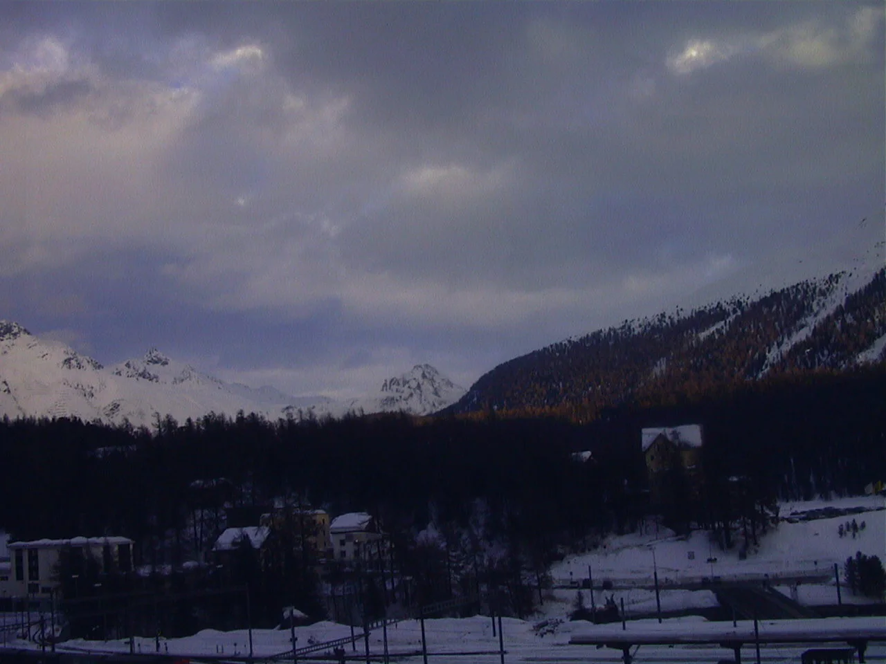 Snowy ground, scattered buildings and wooded slopes lie beneath a thick grey sky and distant white peaks.