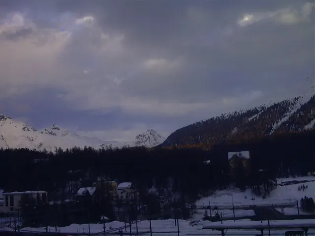 Snowy ground, scattered buildings and wooded slopes lie beneath a thick grey sky and distant white peaks.