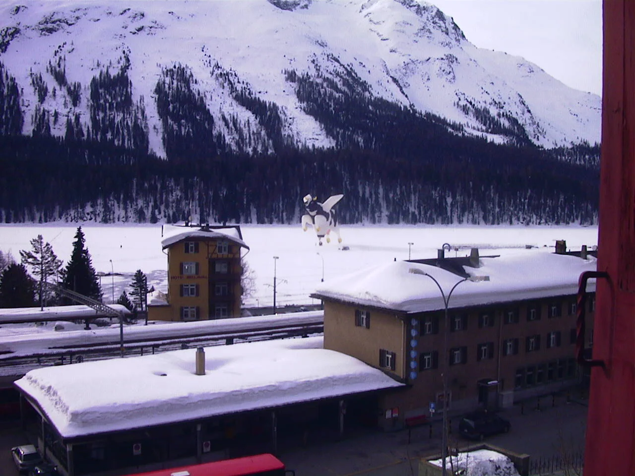 Snow-covered roofs and a clock tower stand below a white mountain slope, with a cow-shaped decoration in the foreground.