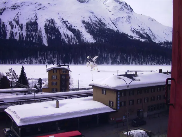 Snow-covered roofs and a clock tower stand below a white mountain slope, with a cow-shaped decoration in the foreground.