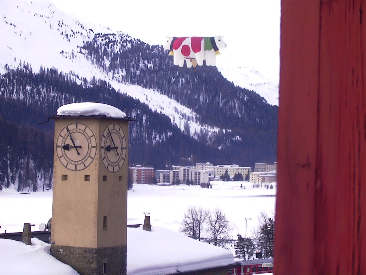 A red window frame borders a view of a clock tower and snowy valley, with a colourful hanging decoration in front.