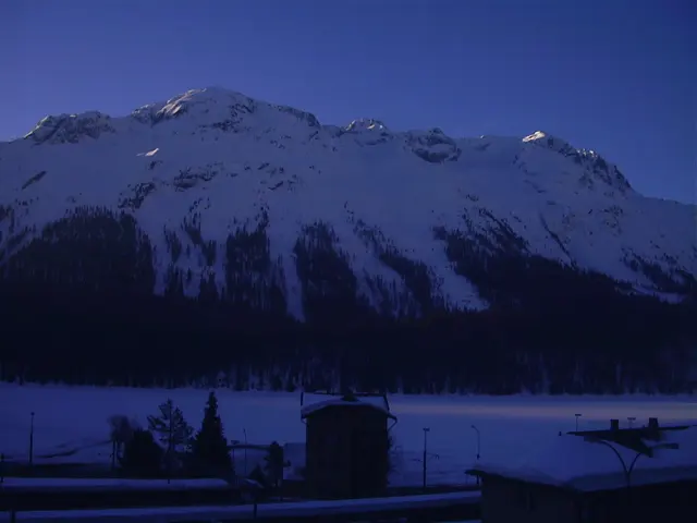 A broad snow-covered mountain rises above dark forest and rooftops beneath a deep blue sky.