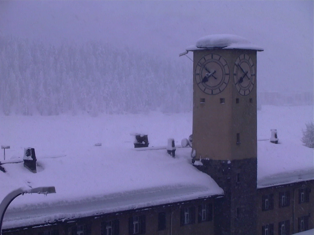A clock tower and low roof are capped with snow against a pale background of trees and snowfall.