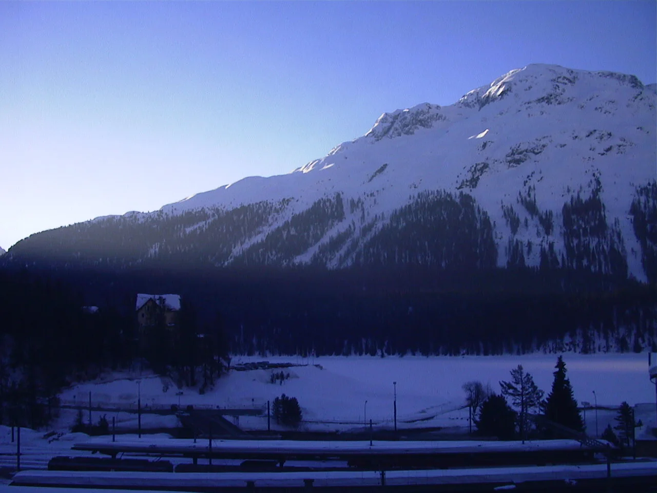 A sunlit snowy mountain rises beyond a dark forest, with snow-covered ground and railway lines below.
