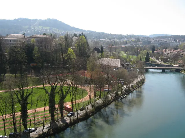 A blue-green river curves beside a tree-lined park and paths, with buildings and a bridge beyond.
