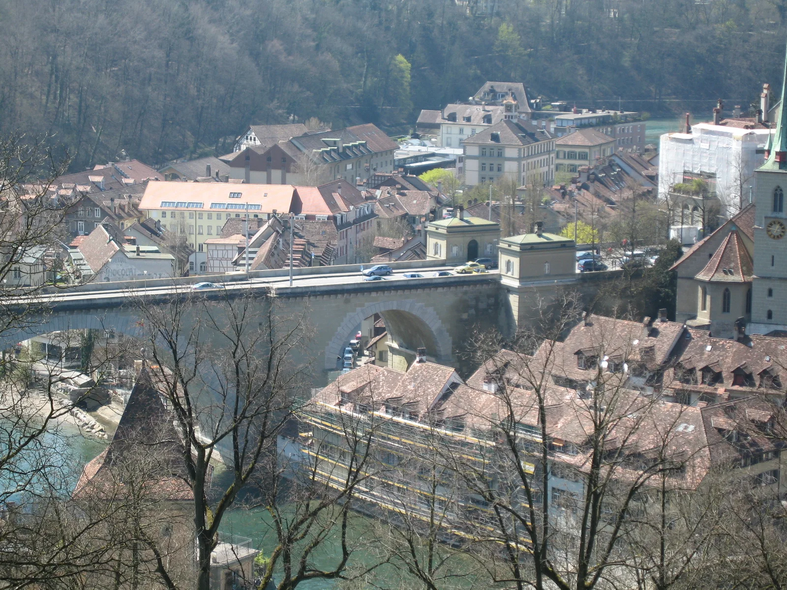 A large stone bridge crosses above clustered red-tiled roofs and a river, with a wooded hillside behind.
