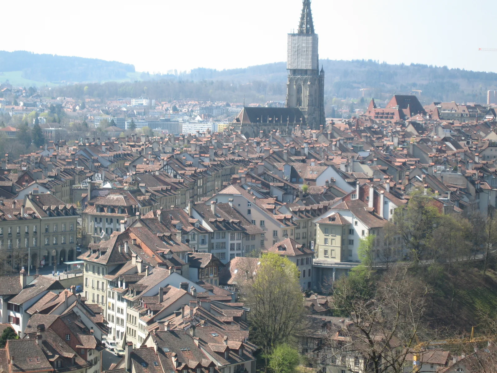 A tall church tower rises over a dense spread of red-tiled rooftops beneath distant wooded hills.