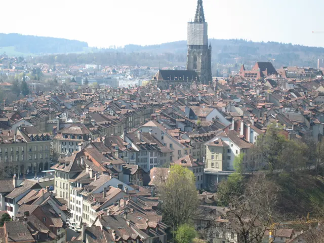 A tall church tower rises over a dense spread of red-tiled rooftops beneath distant wooded hills.