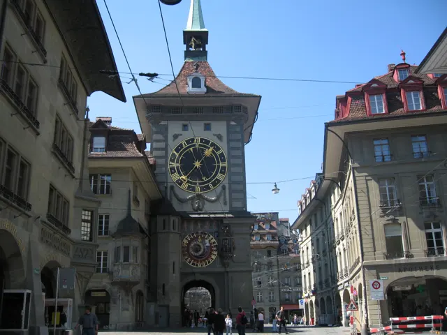 A large clock face and smaller decorative dial mark a stone tower above a narrow street crossed by overhead wires.