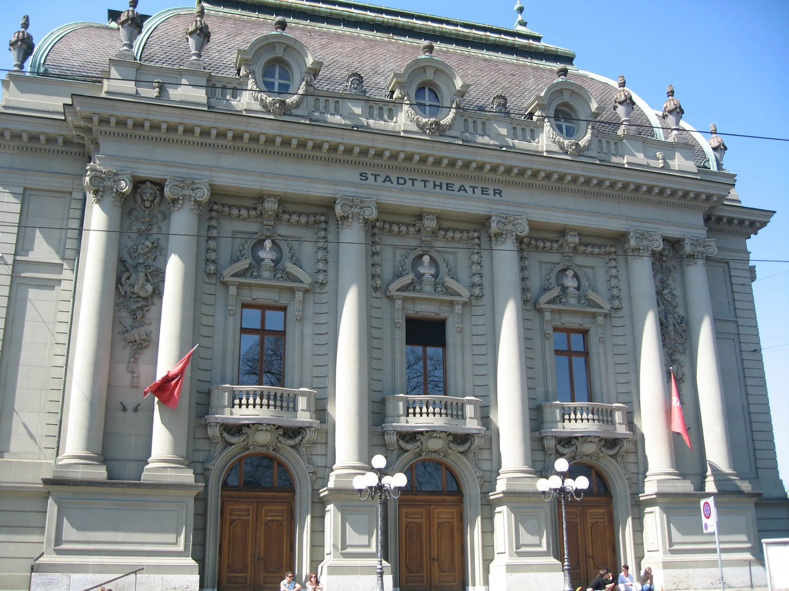 An ornate pale facade with tall columns, arched doors and sculpted roof details stands beneath a clear sky.