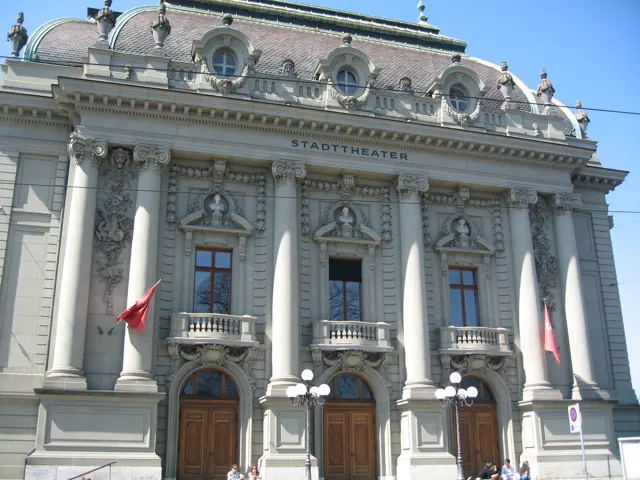 An ornate pale facade with tall columns, arched doors and sculpted roof details stands beneath a clear sky.