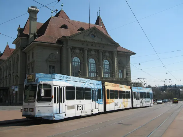 A blue-and-white tram passes a large stone building with a steep red roof beneath a network of overhead wires.