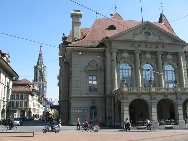 A large stone building with tall arched windows and a red roof faces an open paved square with pedestrians.