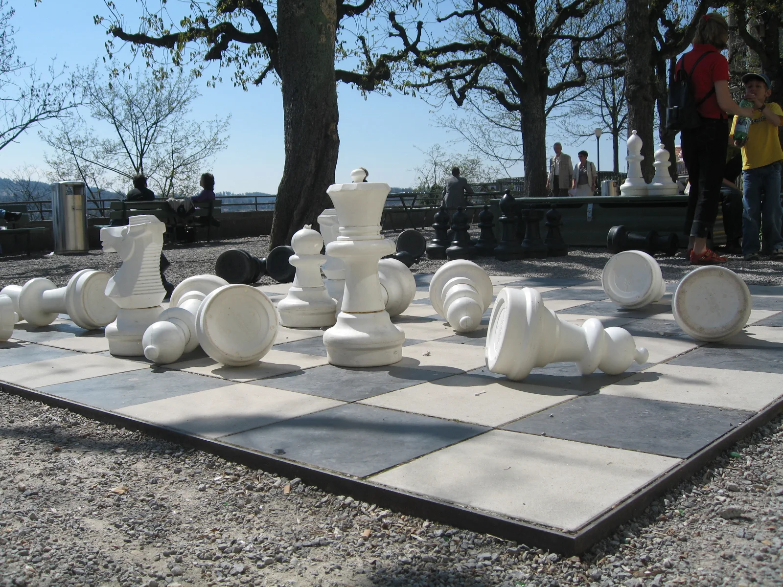 Oversized white chess pieces stand and lie on a large outdoor chessboard beneath trees.