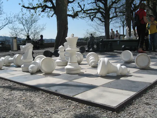 Oversized white chess pieces stand and lie on a large outdoor chessboard beneath trees.