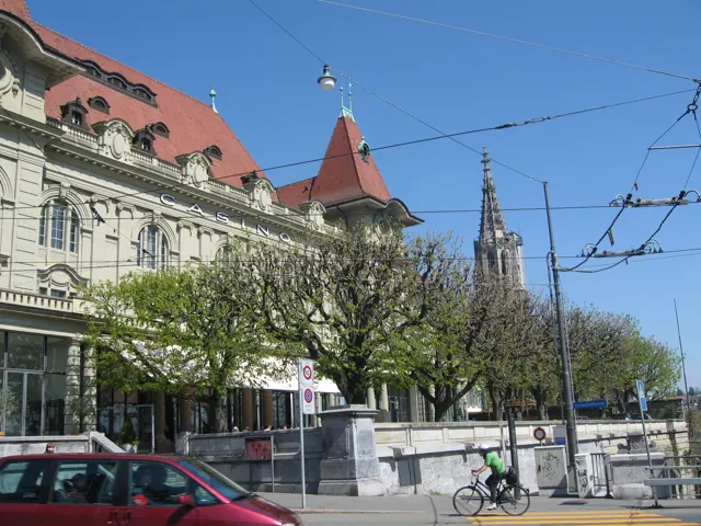 An ornate building with a red roof stands beside street trees, overhead wires and parked vehicles, with a spire beyond.
