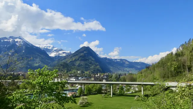 An elevated road bridge crosses a green valley below snow-covered peaks.