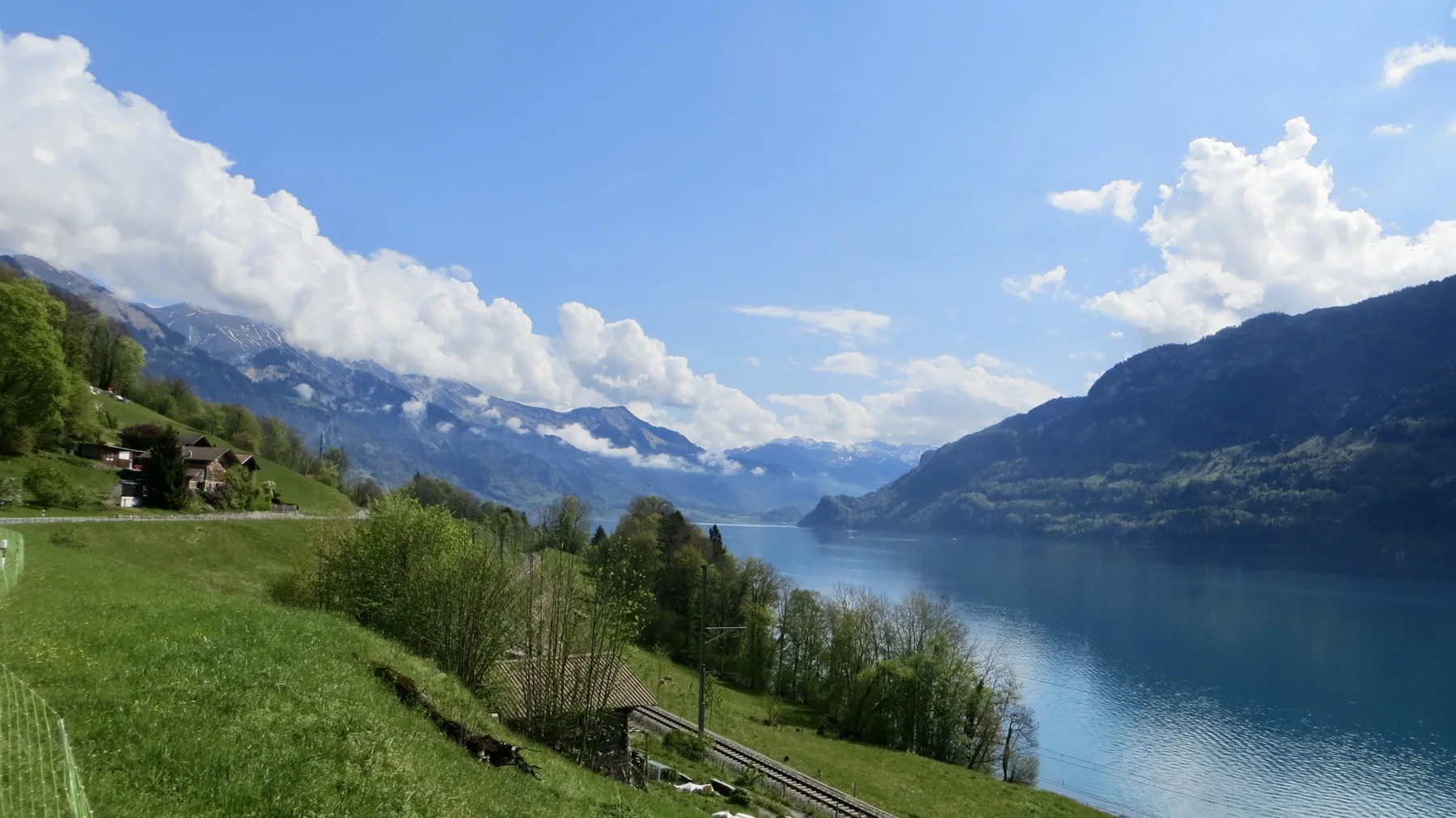 Railway tracks follow a green lakeside slope toward distant mountains.