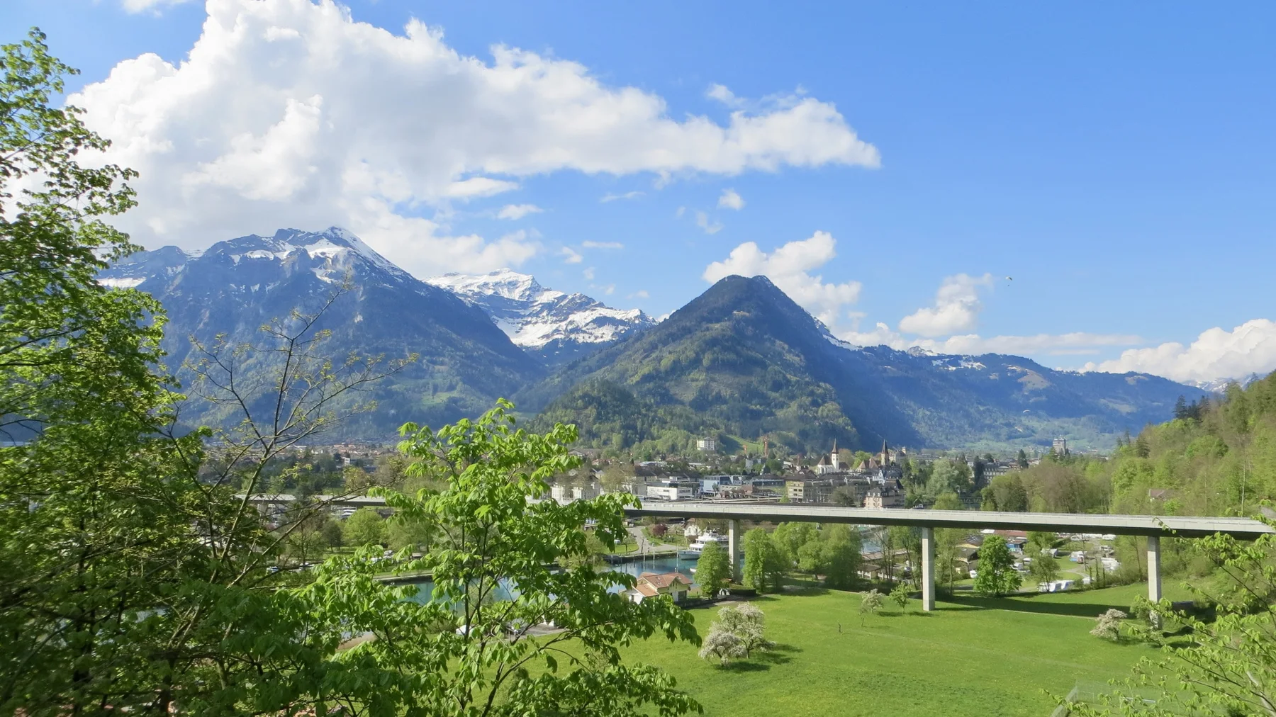 A long road bridge spans a wooded valley with buildings and snowy mountains beyond.