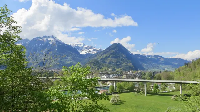 A long road bridge spans a wooded valley with buildings and snowy mountains beyond.