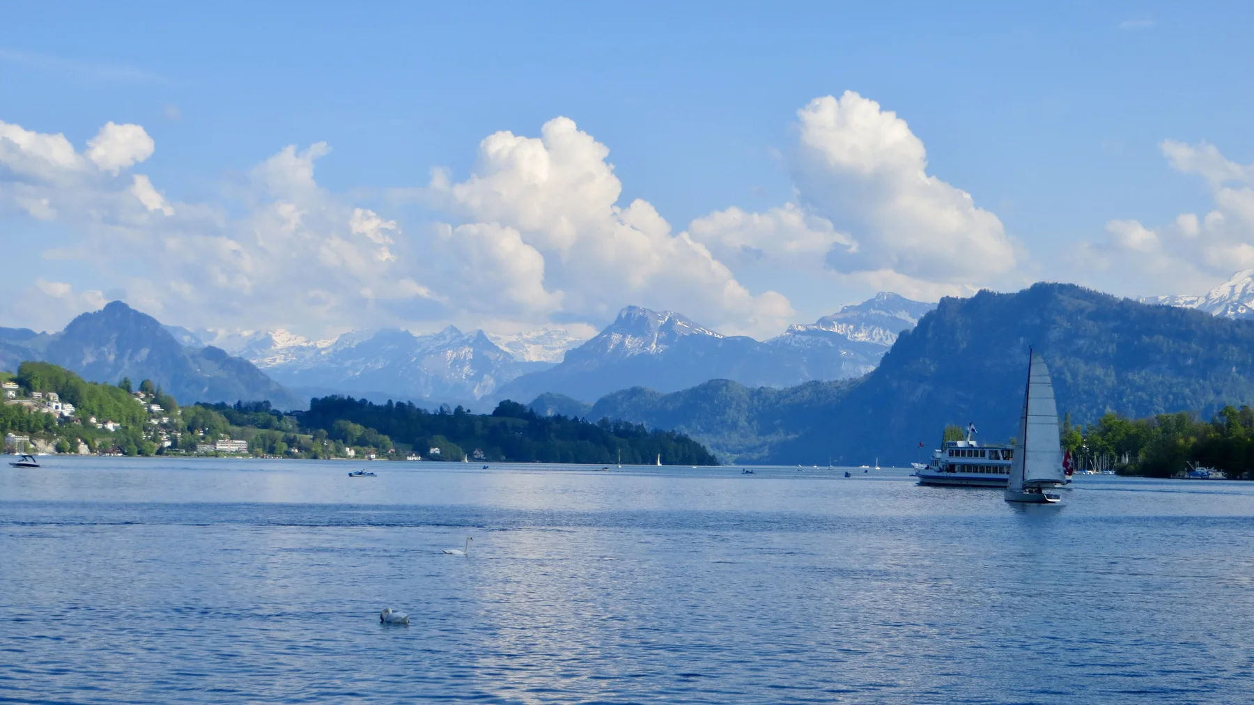 A small sailboat crosses the lake below a shoreline of white houses and mountains.