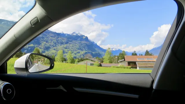A car window and mirror frame a meadow, wooden building and mountains.