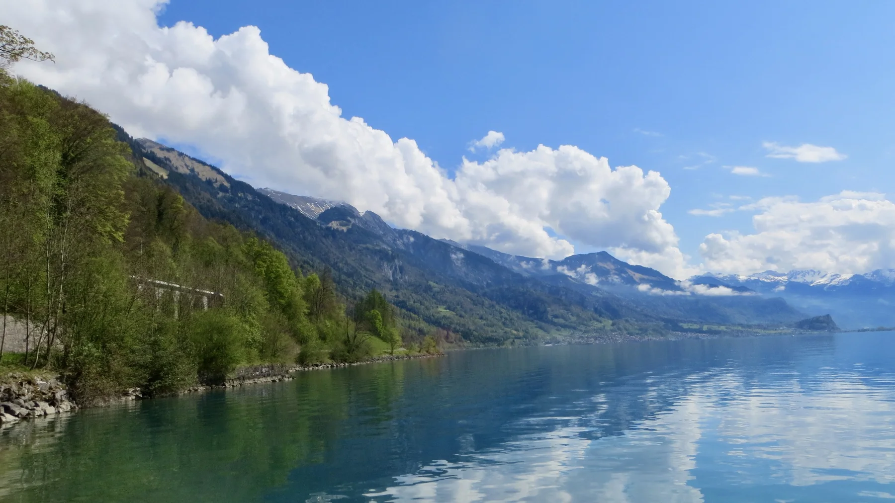 A stone shoreline wall borders a calm lake reflecting wooded, snowy ridges.