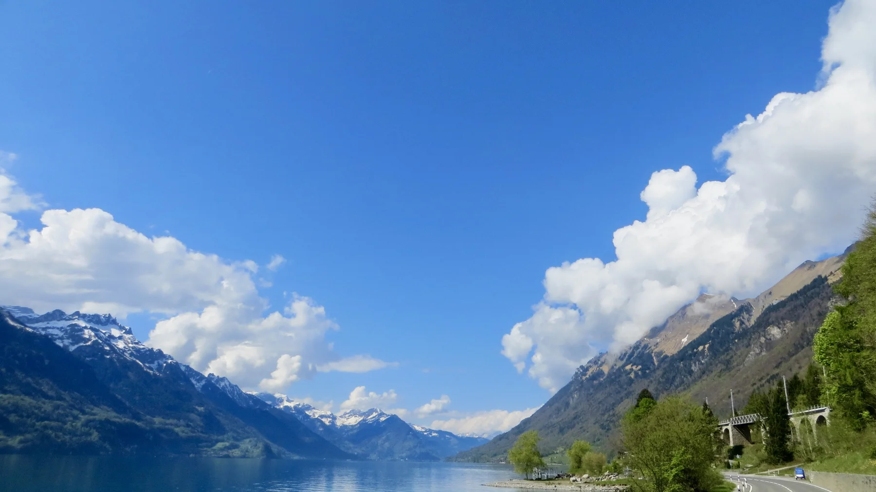 White clouds fill a broad blue sky above a narrow lake between mountains.