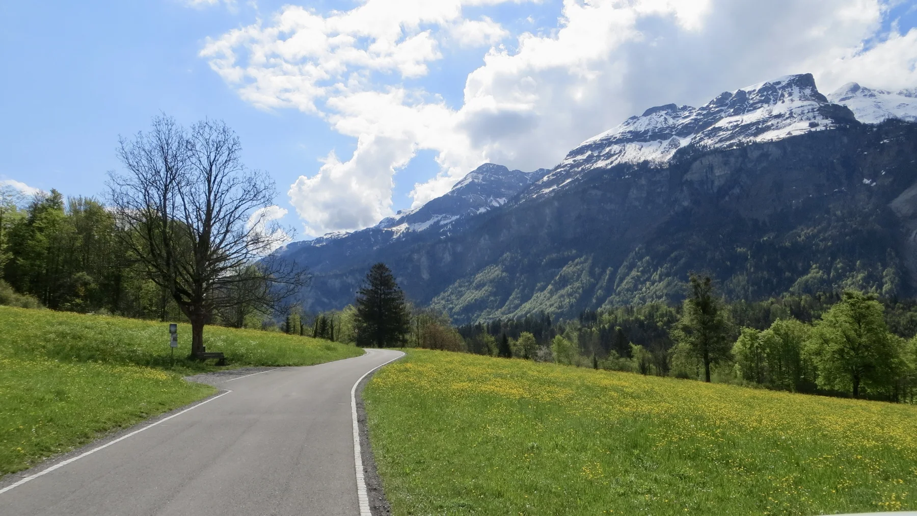 A narrow paved road curves through green meadows below a snowy mountain ridge.