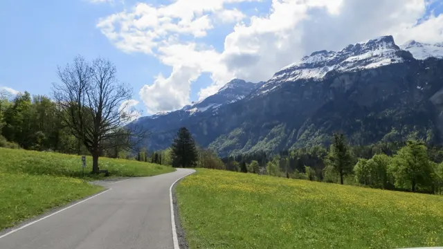 A narrow paved road curves through green meadows below a snowy mountain ridge.
