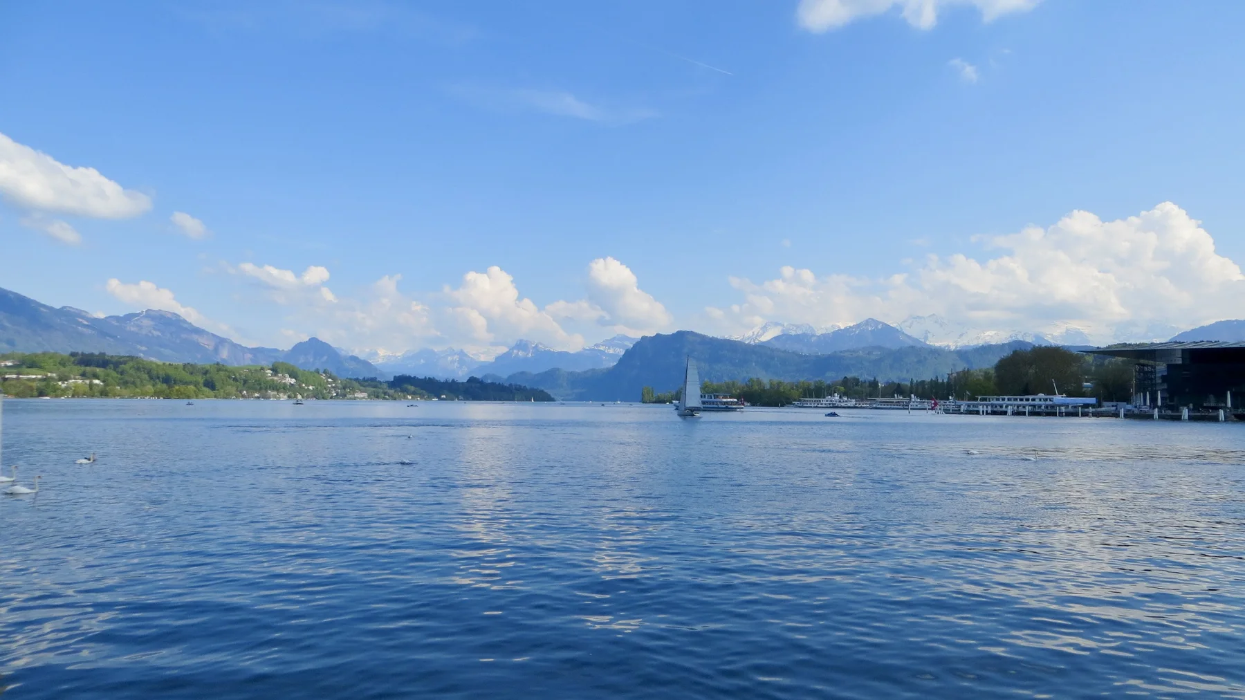 A small sailboat crosses a sunny lake with shoreline buildings and distant mountains.