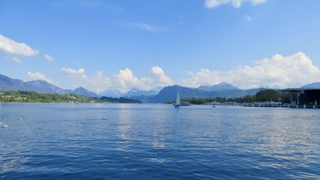 A small sailboat crosses a sunny lake with shoreline buildings and distant mountains.